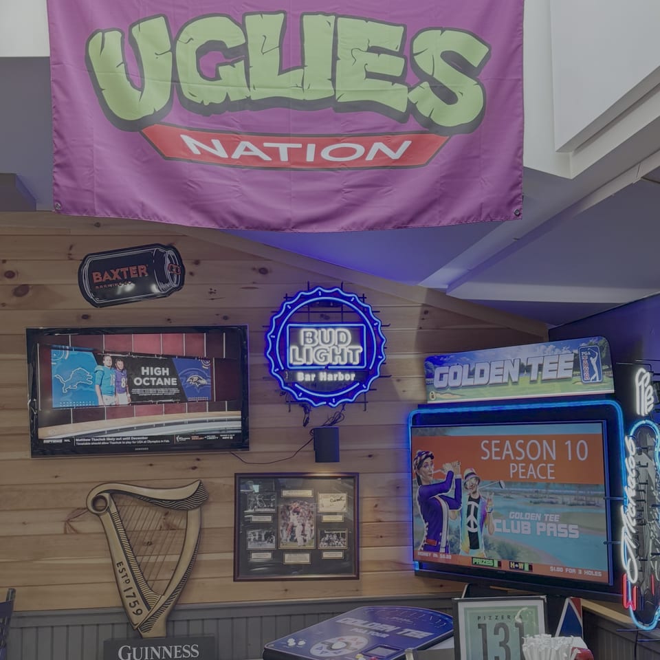 A wall with various signs, including a Bud Light neon sign, a Golden Tee arcade game, and a Guinness harp.