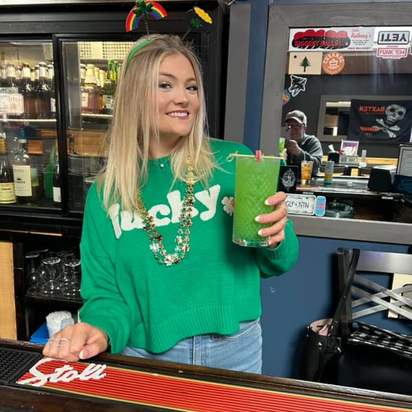 A smiling blonde woman in a green "lucky" sweater holds a green drink at Pizzeria 131's bar.