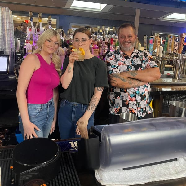 Three bartenders, two women and one man, pose behind a bar with bottles and beer taps.