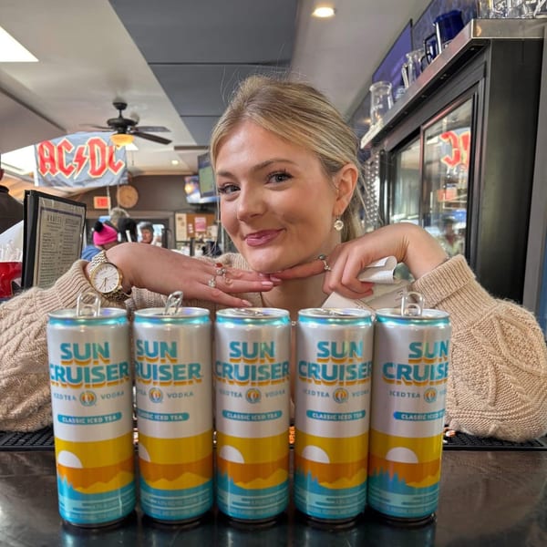 A smiling woman poses behind five cans of Sun Cruiser Iced Tea Vodka at Pizzeria 131.